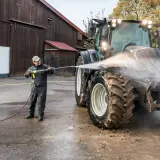 Mężczyzna myje traktor myjką gorącowodną przed stodołą.