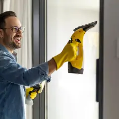 Uśmiechnięty mężczyzna w żółtych rękawiczkach myje okno elektrycznym odkurzaczem do szyb.