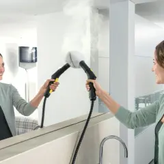 Kobieta czyści lustro parownicą, z której wydobywa się para.