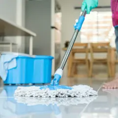 Osoba w rękawiczce myje podłogę mopem. Obok stoi niebieskie wiadro.