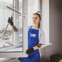Kobieta w niebieskim fartuchu myje okno przy użyciu ściągaczki.