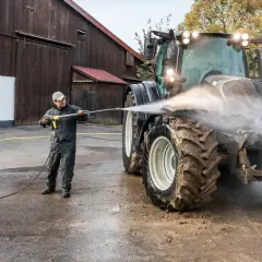 Mężczyzna myje traktor myjką gorącowodną przed stodołą.