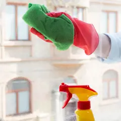 Ręka w czerwonej rękawiczce czyści okno zieloną ściereczką, obok stoi żółto-czerwony spryskiwacz.