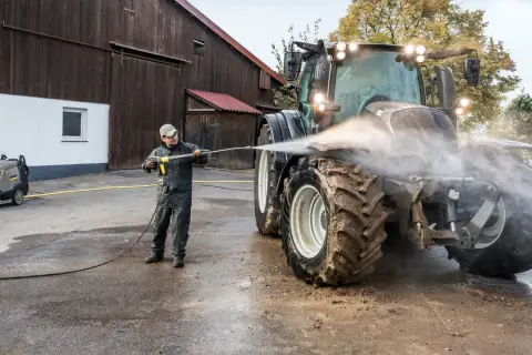 Mężczyzna myje traktor myjką gorącowodną przed stodołą.