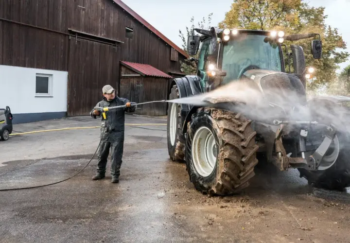 Mężczyzna myje traktor myjką gorącowodną przed stodołą.