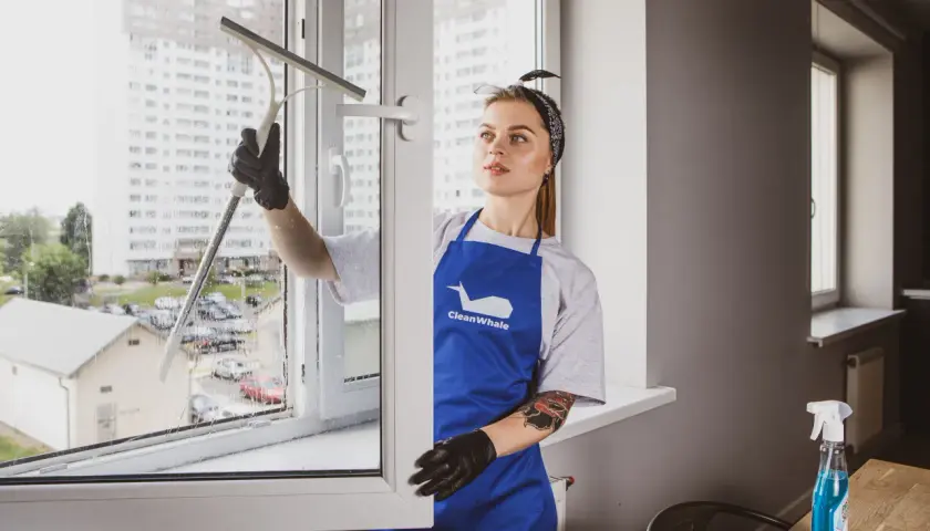 Kobieta w niebieskim fartuchu myje okno przy użyciu ściągaczki.