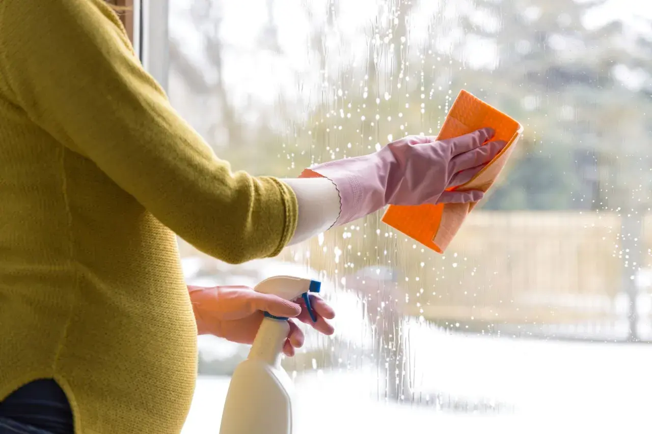 Ciężarna kobieta w rękawiczkach myje okno gąbką i płynem w sprayu.